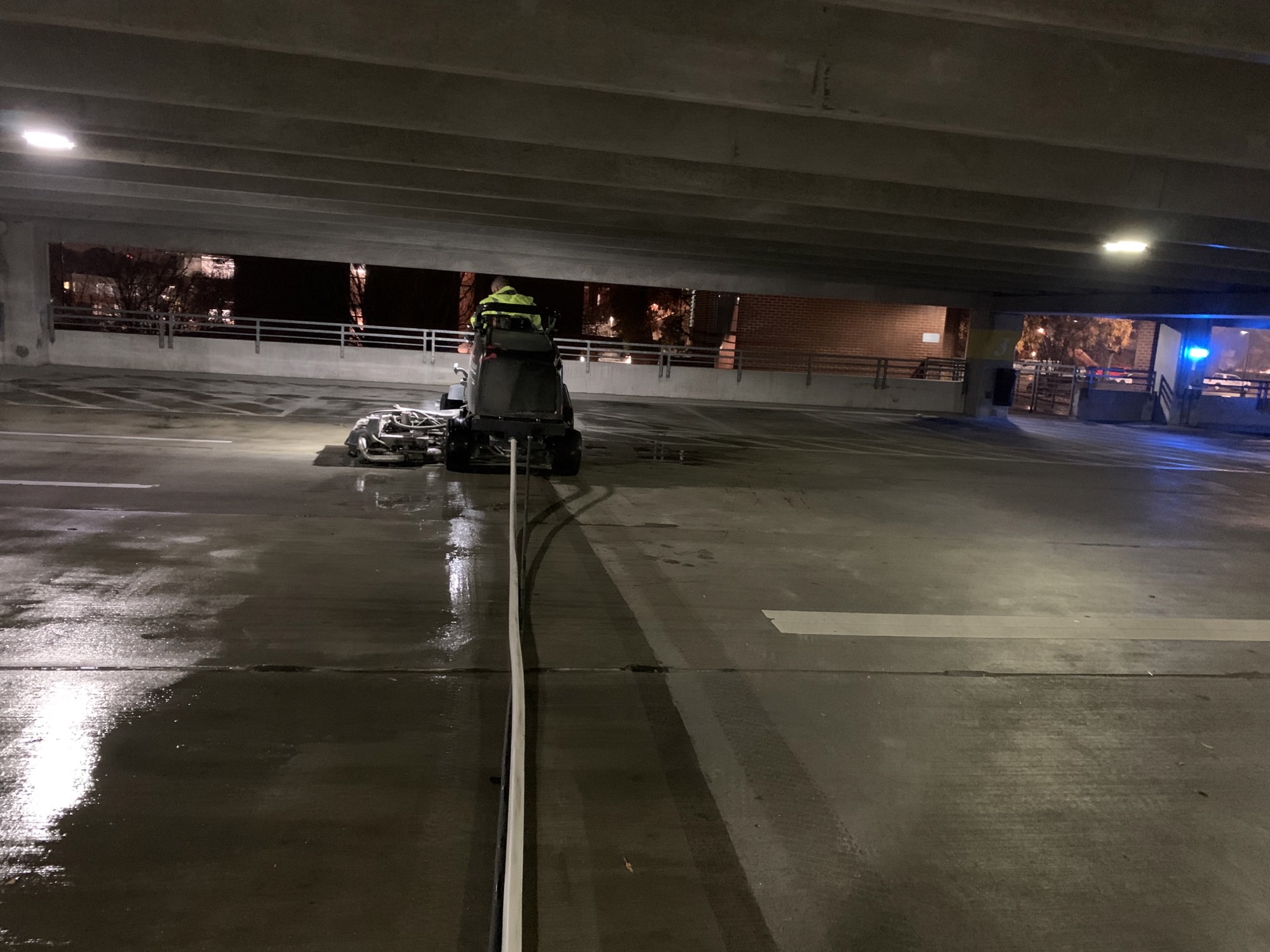 A cleaning machine and worker in a parking garage at night with a long hose on the wet floor.