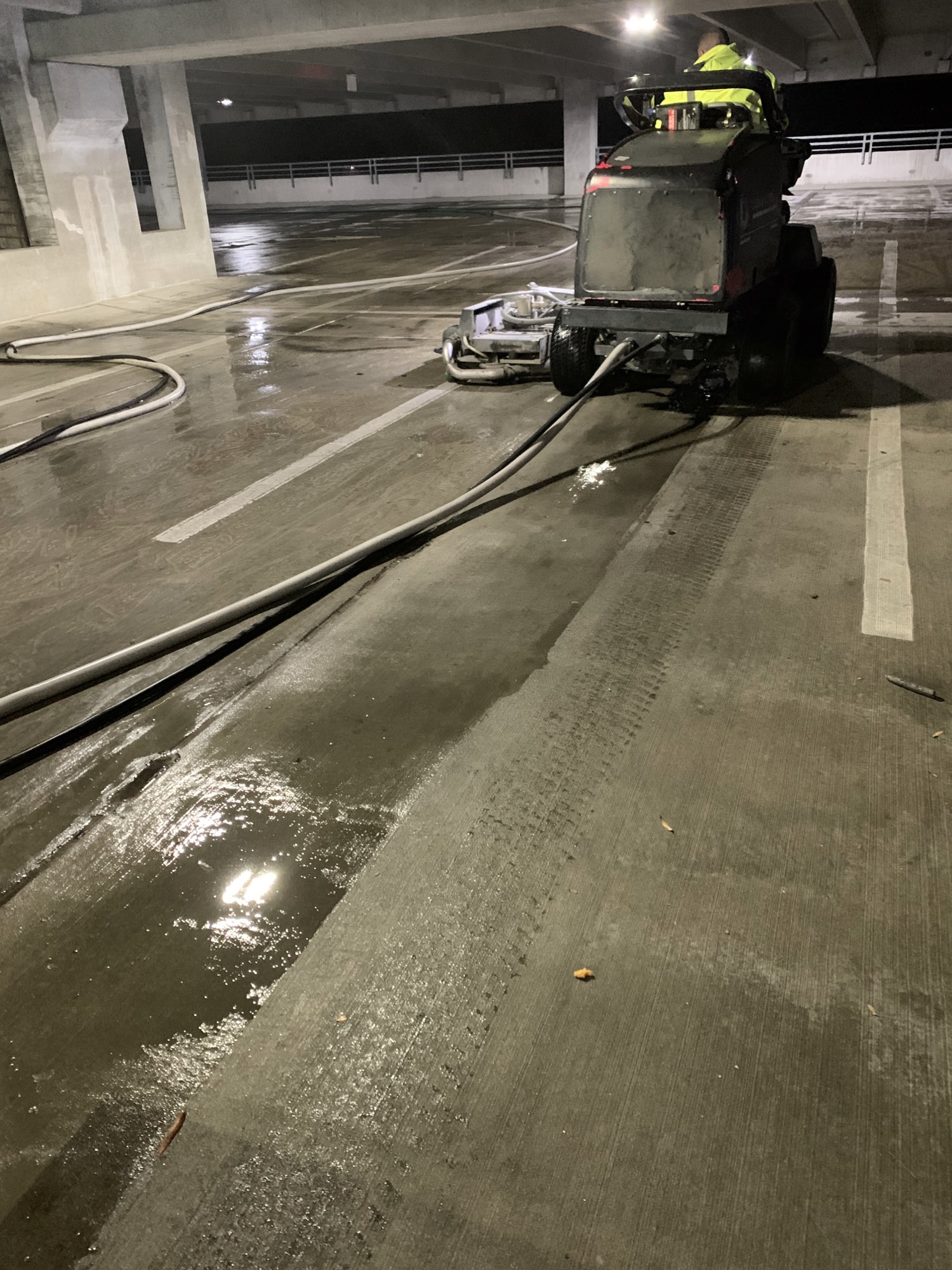 A cleaning machine with an operator cleaning a wet concrete floor in a parking garage.