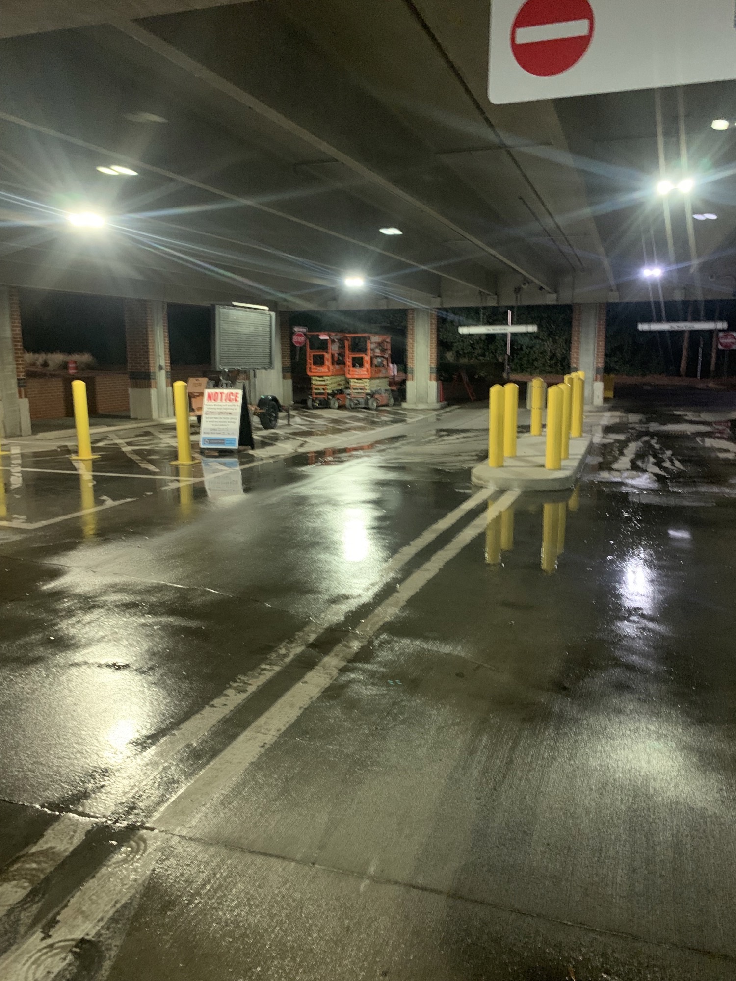 Dimly lit parking garage with wet floors, yellow bollards, and a maintenance lift.