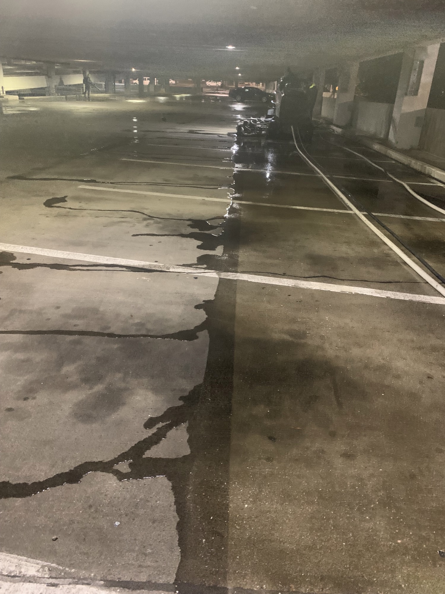 Dimly lit parking garage with wet concrete floors and a cleaning machine with hose.